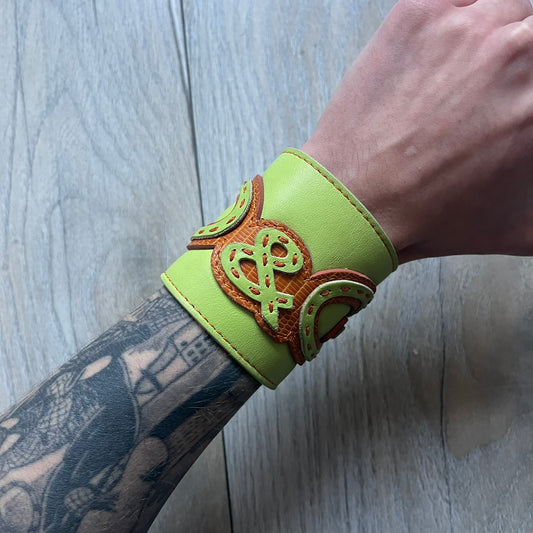 The Dolce &amp; Gabbana Big Green Leather Bracelet is a tall green leather wristband featuring a layered D&amp;G logo along its length - an orange base layer with a green logo on top, detailed with visible orange stitching. The inside has a Dolce &amp; Gabbana tag, and it closes with two metal buttons, offering four total button positions for adjustable sizing.