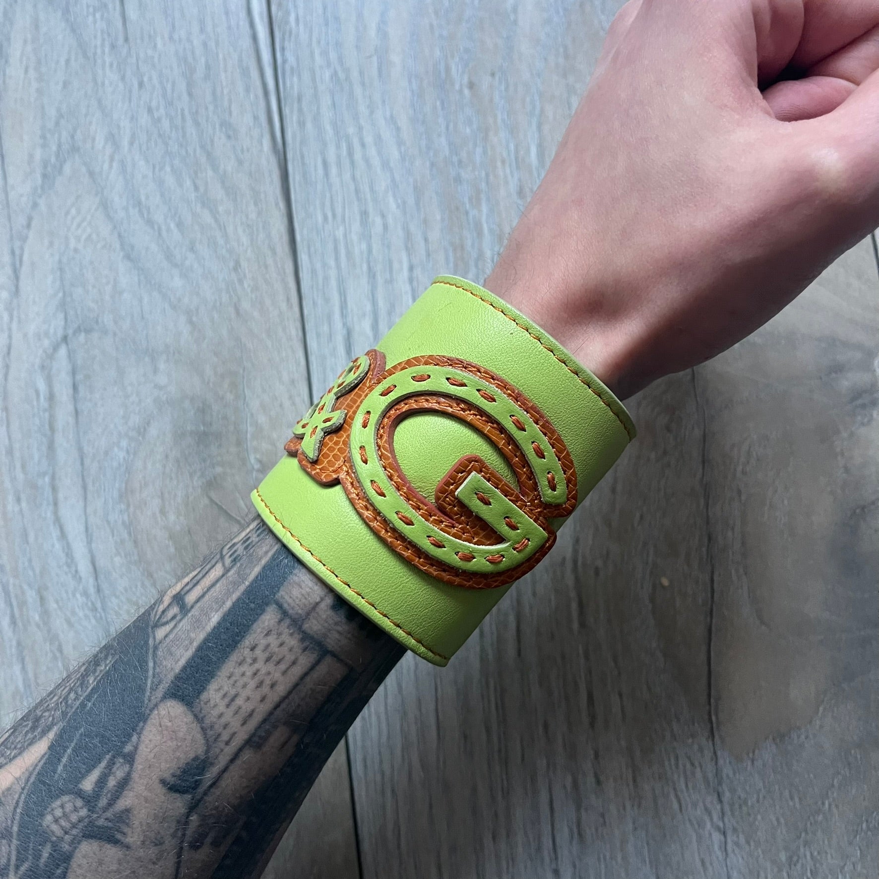 The Dolce &amp; Gabbana Big Green Leather Bracelet is a tall green leather wristband featuring a layered D&amp;G logo along its length - an orange base layer with a green logo on top, detailed with visible orange stitching. The inside has a Dolce &amp; Gabbana tag, and it closes with two metal buttons, offering four total button positions for adjustable sizing.