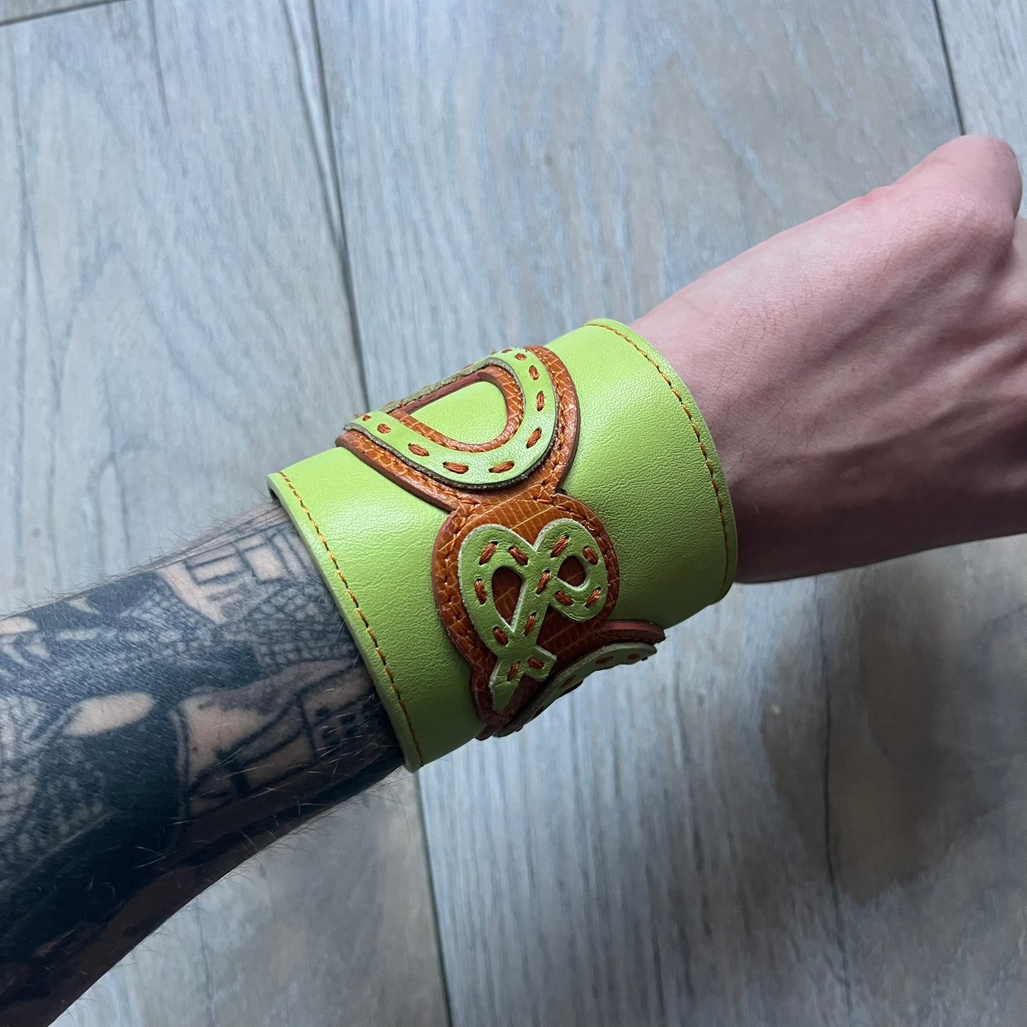 The Dolce &amp; Gabbana Big Green Leather Bracelet is a tall green leather wristband featuring a layered D&amp;G logo along its length - an orange base layer with a green logo on top, detailed with visible orange stitching. The inside has a Dolce &amp; Gabbana tag, and it closes with two metal buttons, offering four total button positions for adjustable sizing.