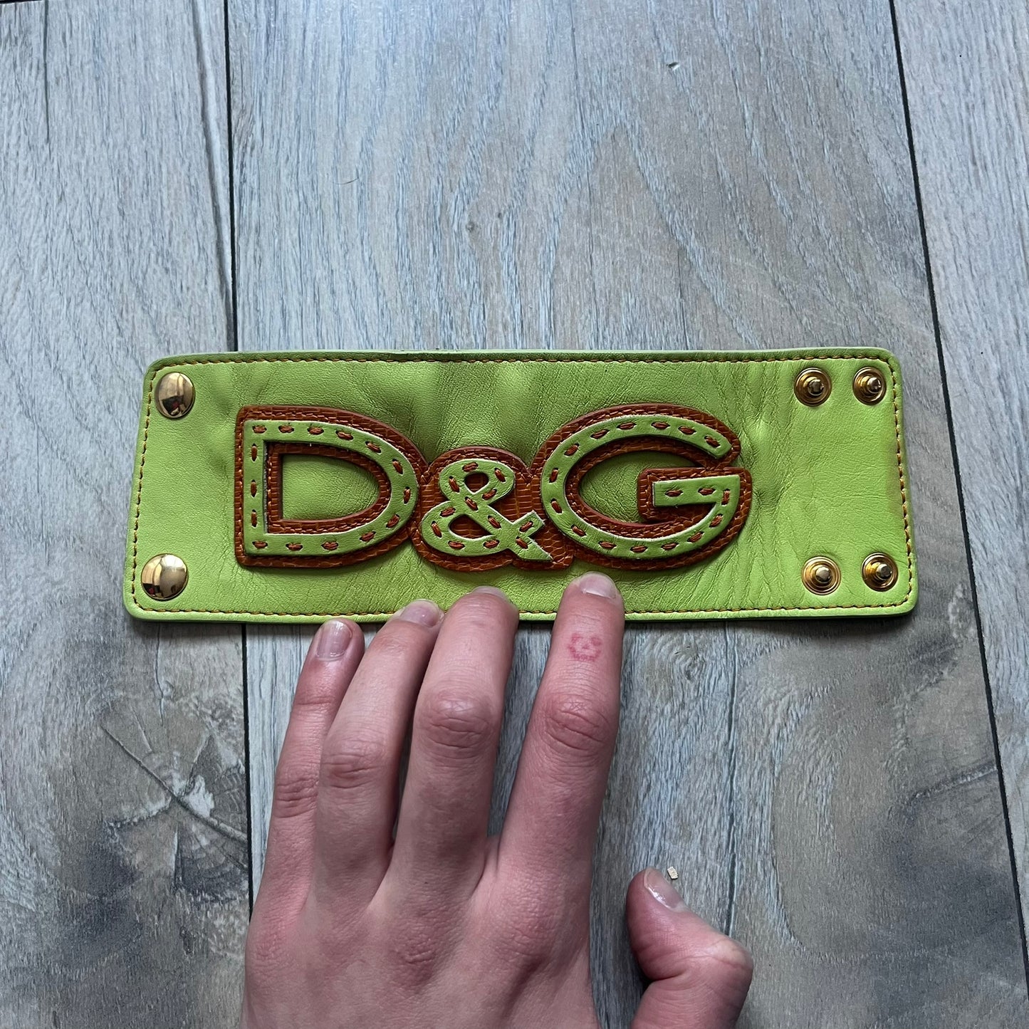 The Dolce &amp; Gabbana Big Green Leather Bracelet is a tall green leather wristband featuring a layered D&amp;G logo along its length - an orange base layer with a green logo on top, detailed with visible orange stitching. The inside has a Dolce &amp; Gabbana tag, and it closes with two metal buttons, offering four total button positions for adjustable sizing.