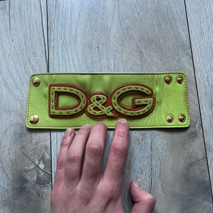 The Dolce &amp; Gabbana Big Green Leather Bracelet is a tall green leather wristband featuring a layered D&amp;G logo along its length - an orange base layer with a green logo on top, detailed with visible orange stitching. The inside has a Dolce &amp; Gabbana tag, and it closes with two metal buttons, offering four total button positions for adjustable sizing.