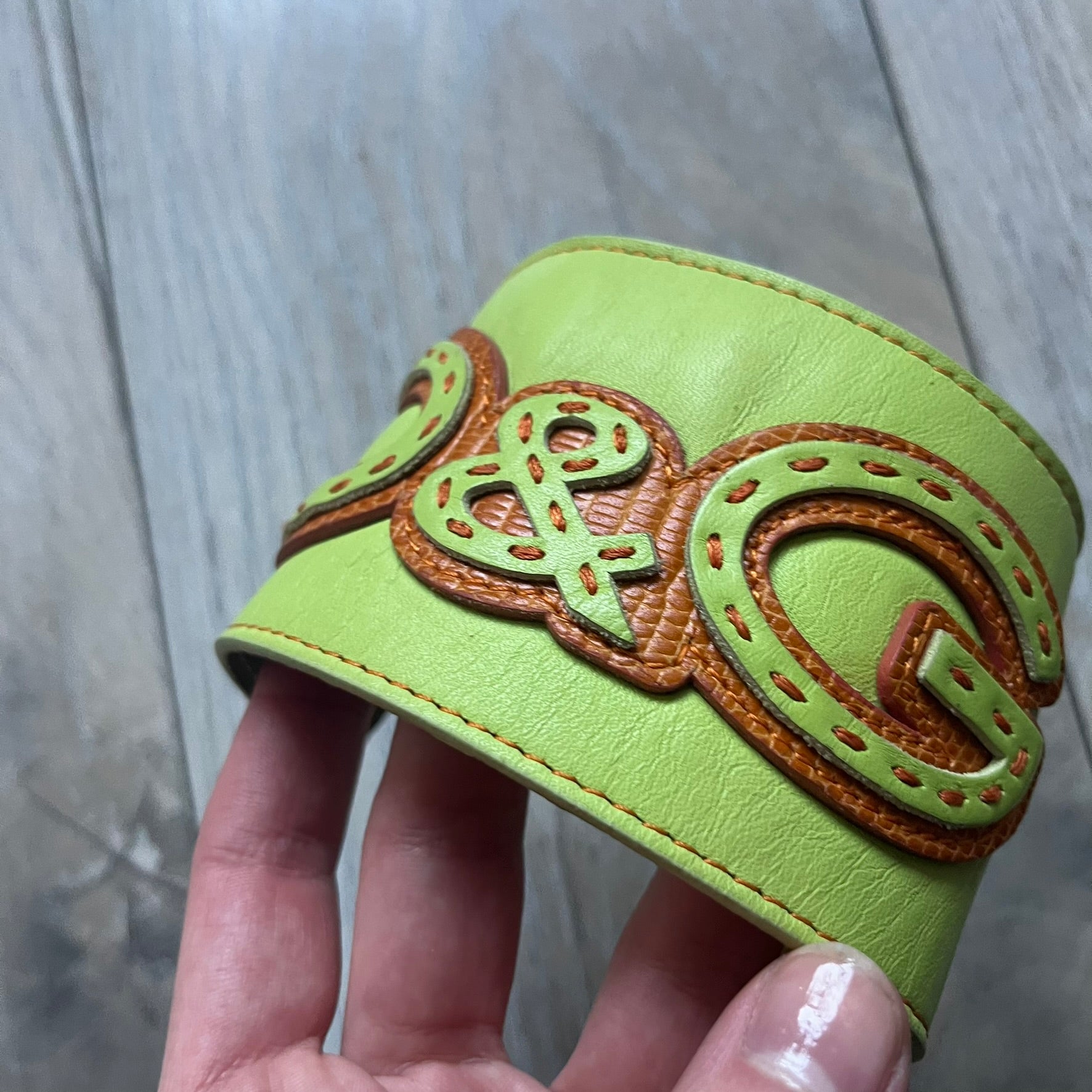 The Dolce &amp; Gabbana Big Green Leather Bracelet is a tall green leather wristband featuring a layered D&amp;G logo along its length - an orange base layer with a green logo on top, detailed with visible orange stitching. The inside has a Dolce &amp; Gabbana tag, and it closes with two metal buttons, offering four total button positions for adjustable sizing.