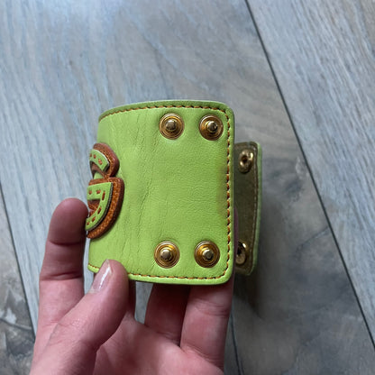 The Dolce &amp; Gabbana Big Green Leather Bracelet is a tall green leather wristband featuring a layered D&amp;G logo along its length - an orange base layer with a green logo on top, detailed with visible orange stitching. The inside has a Dolce &amp; Gabbana tag, and it closes with two metal buttons, offering four total button positions for adjustable sizing.