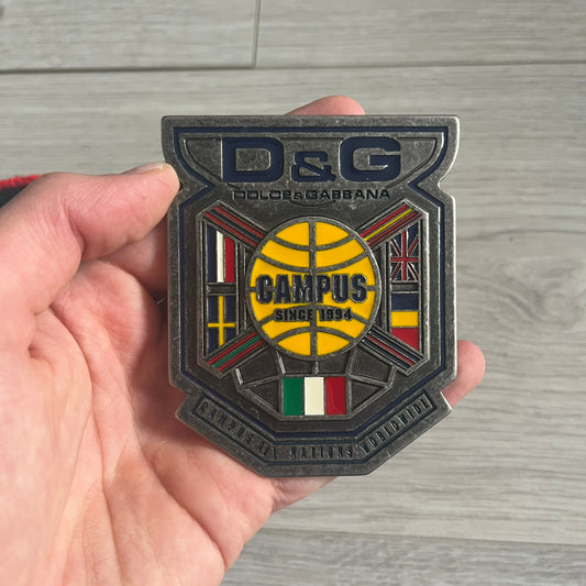 Dolce &amp; Gabbana "Campus Since 1994" Material Belt features a multicolor cotton webbing strap in red, navy, green, and yellow with leather-tipped ends. The statement metal buckle displays a crest design with D&amp;G branding, a central “Campus Since 1994” motif, and multiple national flags. A sporty accessory that combines vintage with luxury detail.
