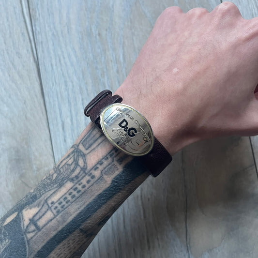 Dolce & Gabbana Champagne Leather Bracelet DJ1097 is a brown leather bracelet designed to resemble a miniature belt, featuring multiple adjustment holes. The silver-tone metal plate is shaped like a D&G champagne label and displays a black D&G logo, along with engraved “Glamorous Champagne 1994 Alc. 12% by Vol.” text. The back of the plate features a large, shallow engraved D&G logo.