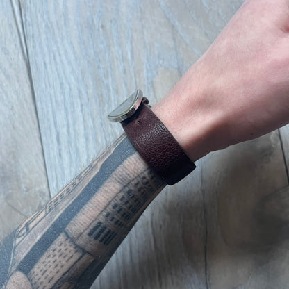 Dolce & Gabbana Champagne Leather Bracelet DJ1097 is a brown leather bracelet designed to resemble a miniature belt, featuring multiple adjustment holes. The silver-tone metal plate is shaped like a D&G champagne label and displays a black D&G logo, along with engraved “Glamorous Champagne 1994 Alc. 12% by Vol.” text. The back of the plate features a large, shallow engraved D&G logo.