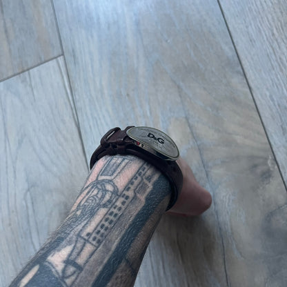 Dolce & Gabbana Champagne Leather Bracelet DJ1097 is a brown leather bracelet designed to resemble a miniature belt, featuring multiple adjustment holes. The silver-tone metal plate is shaped like a D&G champagne label and displays a black D&G logo, along with engraved “Glamorous Champagne 1994 Alc. 12% by Vol.” text. The back of the plate features a large, shallow engraved D&G logo.