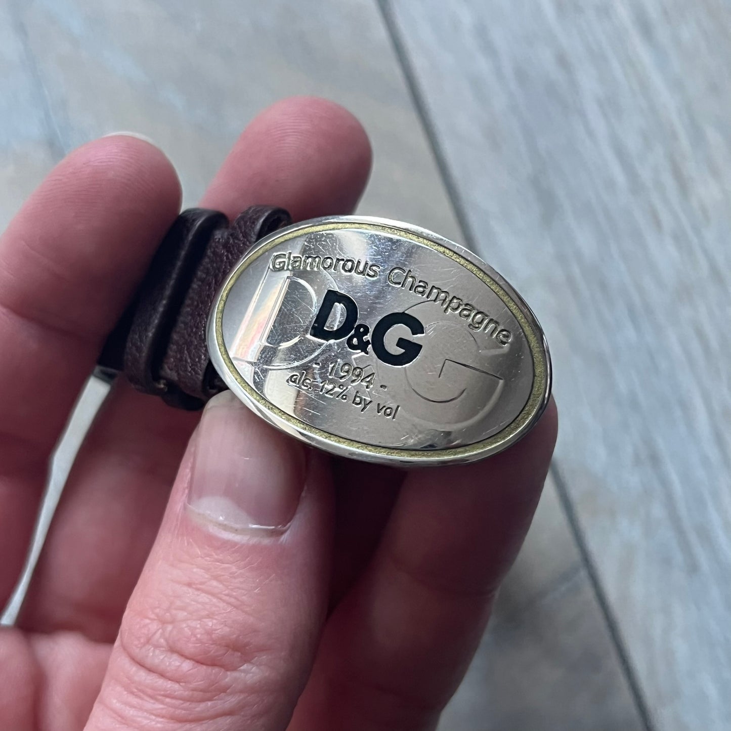 Dolce & Gabbana Champagne Leather Bracelet DJ1097 is a brown leather bracelet designed to resemble a miniature belt, featuring multiple adjustment holes. The silver-tone metal plate is shaped like a D&G champagne label and displays a black D&G logo, along with engraved “Glamorous Champagne 1994 Alc. 12% by Vol.” text. The back of the plate features a large, shallow engraved D&G logo.