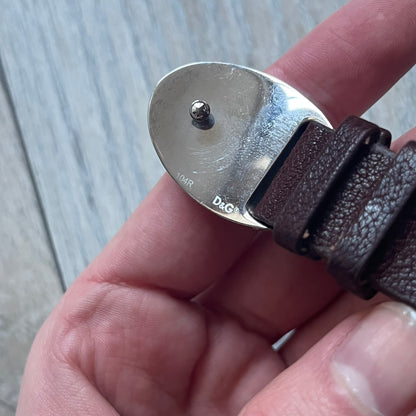 Dolce & Gabbana Champagne Leather Bracelet DJ1097 is a brown leather bracelet designed to resemble a miniature belt, featuring multiple adjustment holes. The silver-tone metal plate is shaped like a D&G champagne label and displays a black D&G logo, along with engraved “Glamorous Champagne 1994 Alc. 12% by Vol.” text. The back of the plate features a large, shallow engraved D&G logo.