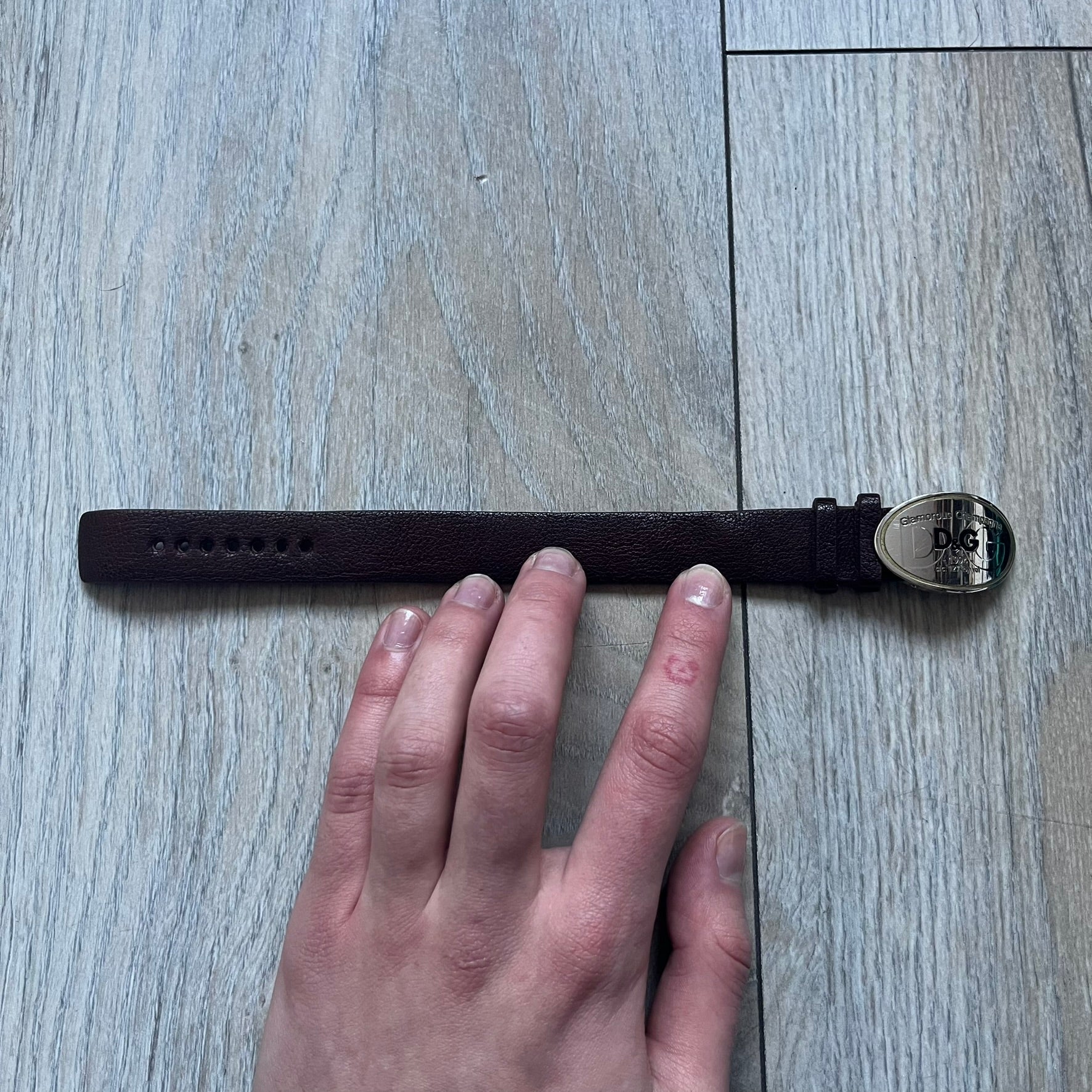 Dolce & Gabbana Champagne Leather Bracelet DJ1097 is a brown leather bracelet designed to resemble a miniature belt, featuring multiple adjustment holes. The silver-tone metal plate is shaped like a D&G champagne label and displays a black D&G logo, along with engraved “Glamorous Champagne 1994 Alc. 12% by Vol.” text. The back of the plate features a large, shallow engraved D&G logo.