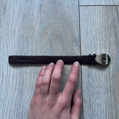 Dolce & Gabbana Champagne Leather Bracelet DJ1097 is a brown leather bracelet designed to resemble a miniature belt, featuring multiple adjustment holes. The silver-tone metal plate is shaped like a D&G champagne label and displays a black D&G logo, along with engraved “Glamorous Champagne 1994 Alc. 12% by Vol.” text. The back of the plate features a large, shallow engraved D&G logo.