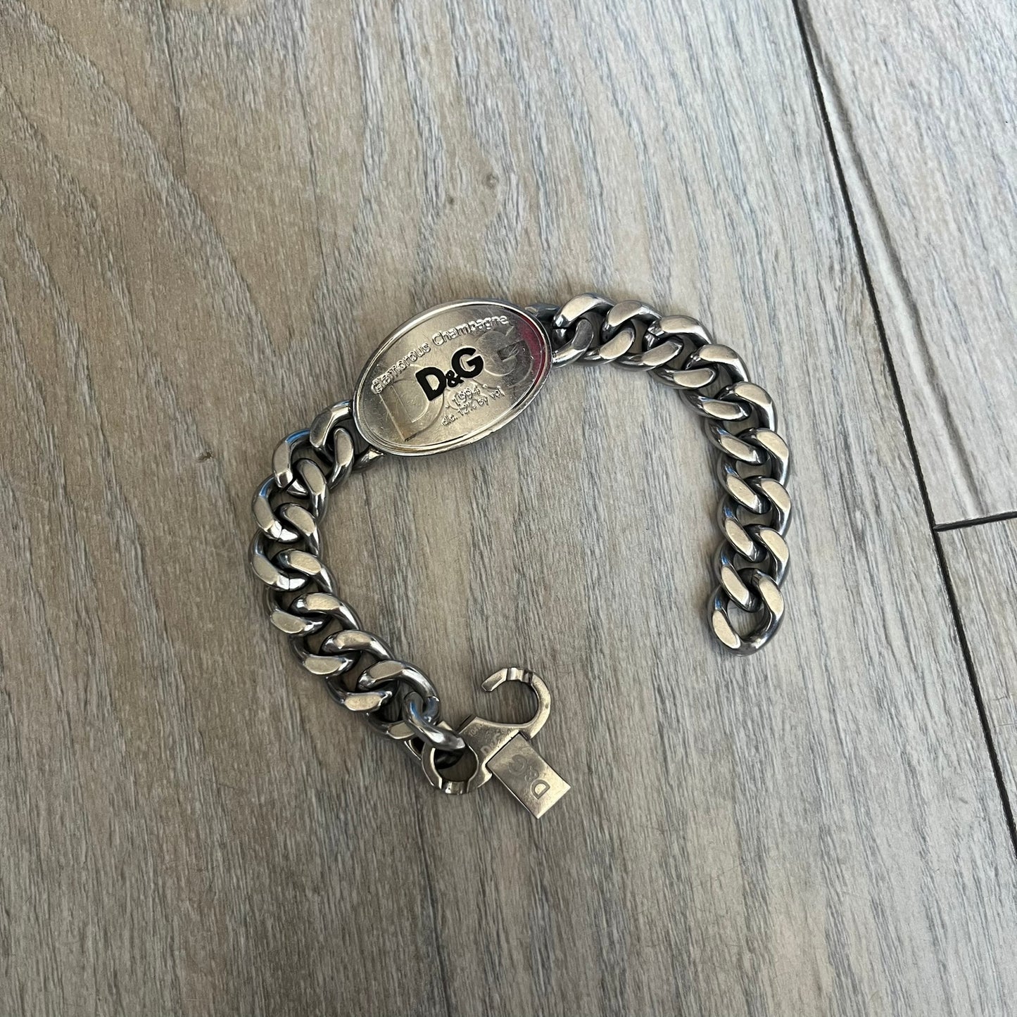 Dolce &amp; Gabbana Champagne Bracelet DJ1092 is a stainless steel bracelet. The silver-tone metal plate is shaped like a D&amp;G champagne label and displays a black D&amp;G logo, along with engraved “Glamorous Champagne 1994 Alc. 12% by Vol.” text. The back of the plate features a large, shallow engraved D&amp;G logo. Retailed at 125 usd in 2000's.
