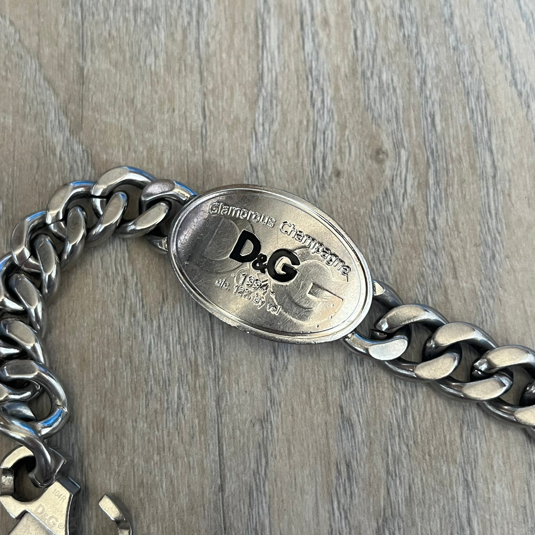 Dolce &amp; Gabbana Champagne Bracelet DJ1092 is a stainless steel bracelet. The silver-tone metal plate is shaped like a D&amp;G champagne label and displays a black D&amp;G logo, along with engraved “Glamorous Champagne 1994 Alc. 12% by Vol.” text. The back of the plate features a large, shallow engraved D&amp;G logo. Retailed at 125 usd in 2000's.