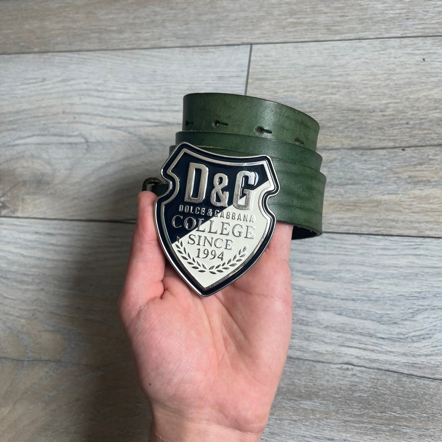 Dolce &amp; Gabbana “College Since 1994” belt featuring a big crest-shaped buckle. The silver text overlays a beige-and-black diagonally split crest, paired with a soft green leather strap in a washed style.