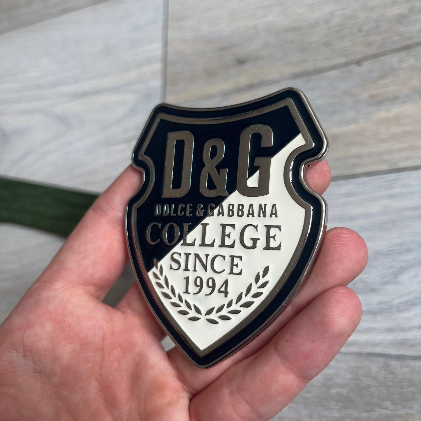 Dolce &amp; Gabbana “College Since 1994” belt featuring a big crest-shaped buckle. The silver text overlays a beige-and-black diagonally split crest, paired with a soft green leather strap in a washed style.