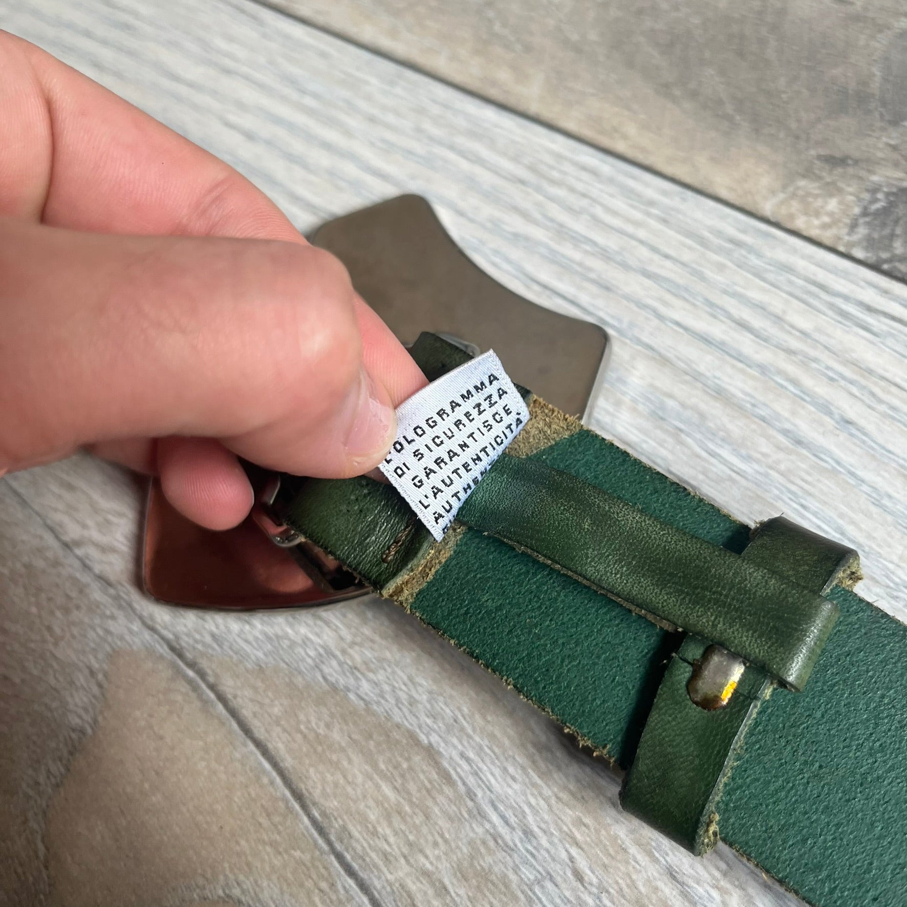 Dolce &amp; Gabbana “College Since 1994” belt featuring a big crest-shaped buckle. The silver text overlays a beige-and-black diagonally split crest, paired with a soft green leather strap in a washed style.