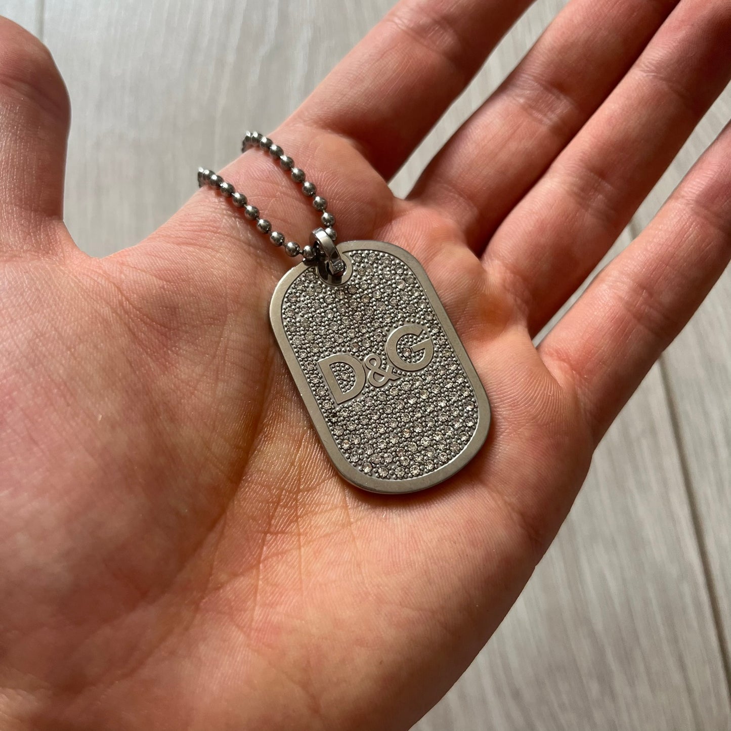 Silver dog tag with 'D&G' logo held in a hand against a light background
