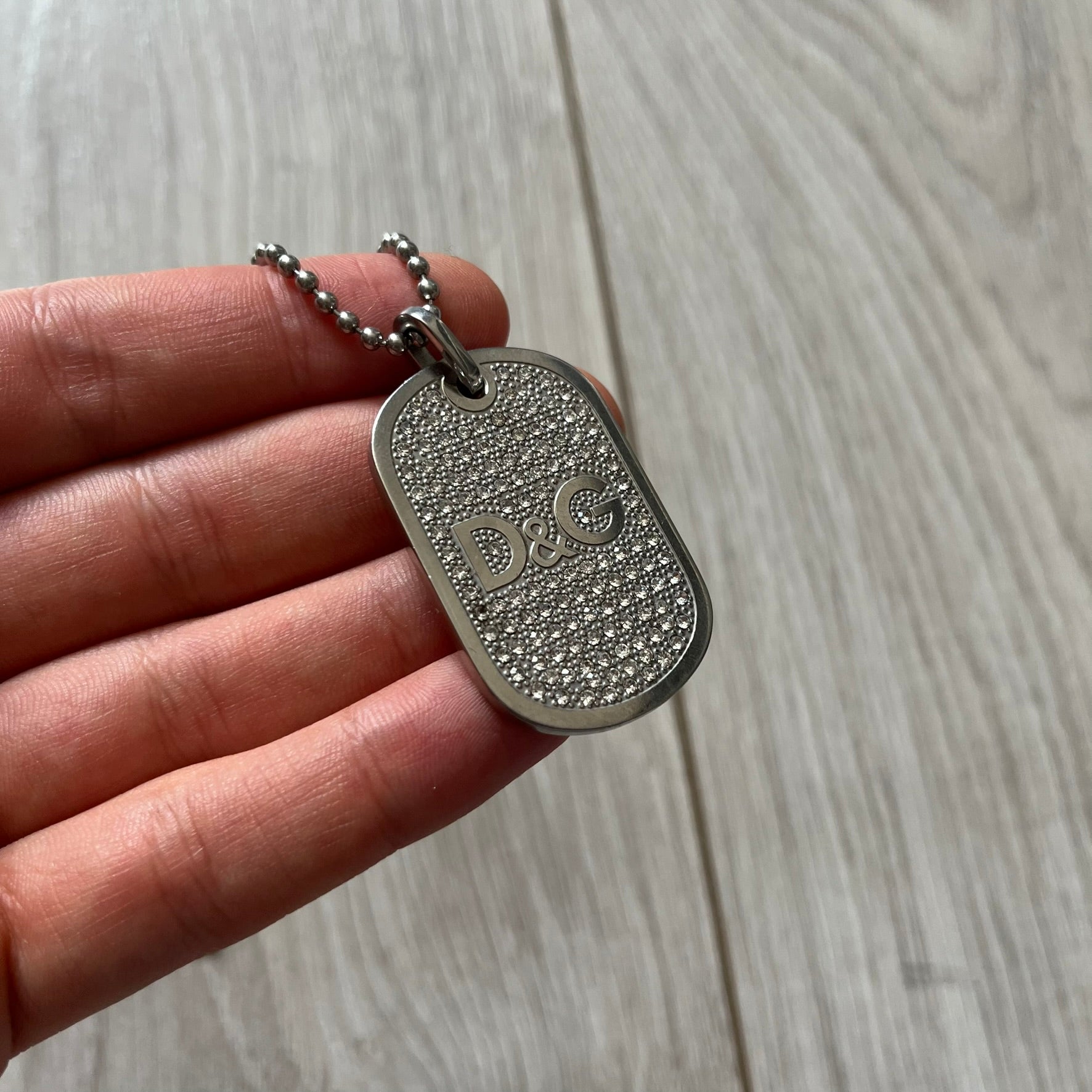Silver dog tag with 'D&G' logo held by a hand on a wooden surface
