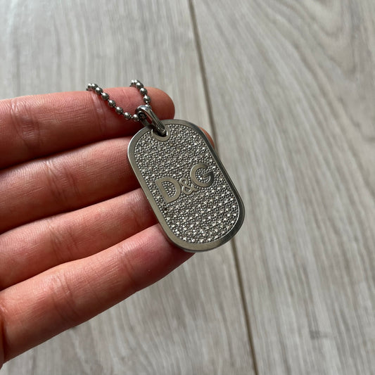 Silver dog tag with 'D&G' logo held by a hand on a wooden surface