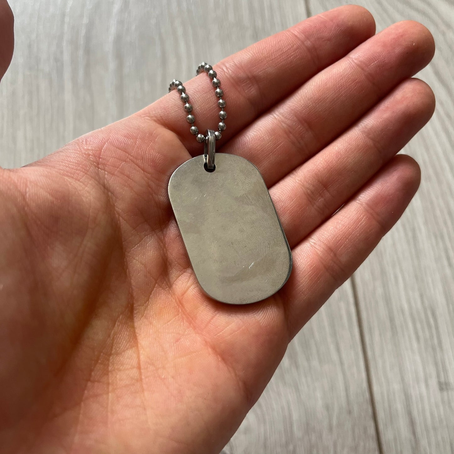 Hand holding a silver dog tag necklace on a wooden surface