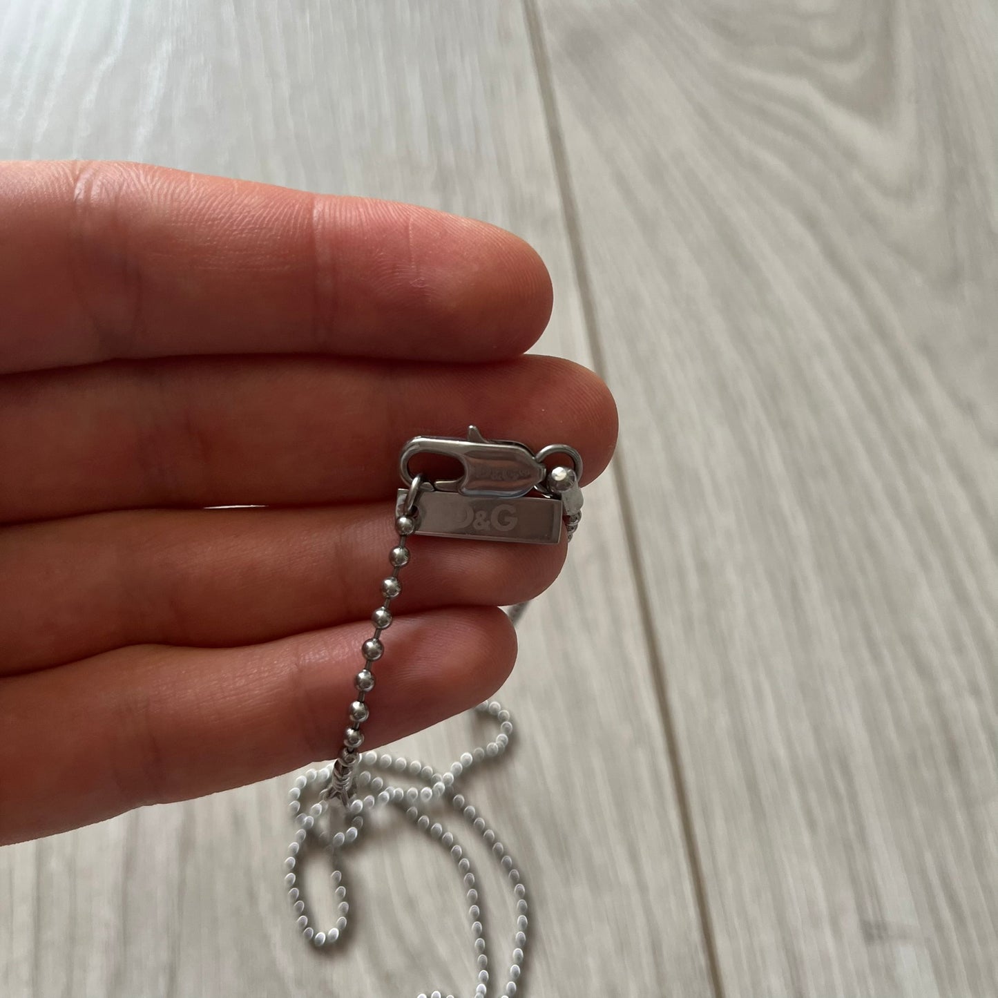 Hand holding a silver chain with a D&G tag against a light wooden surface