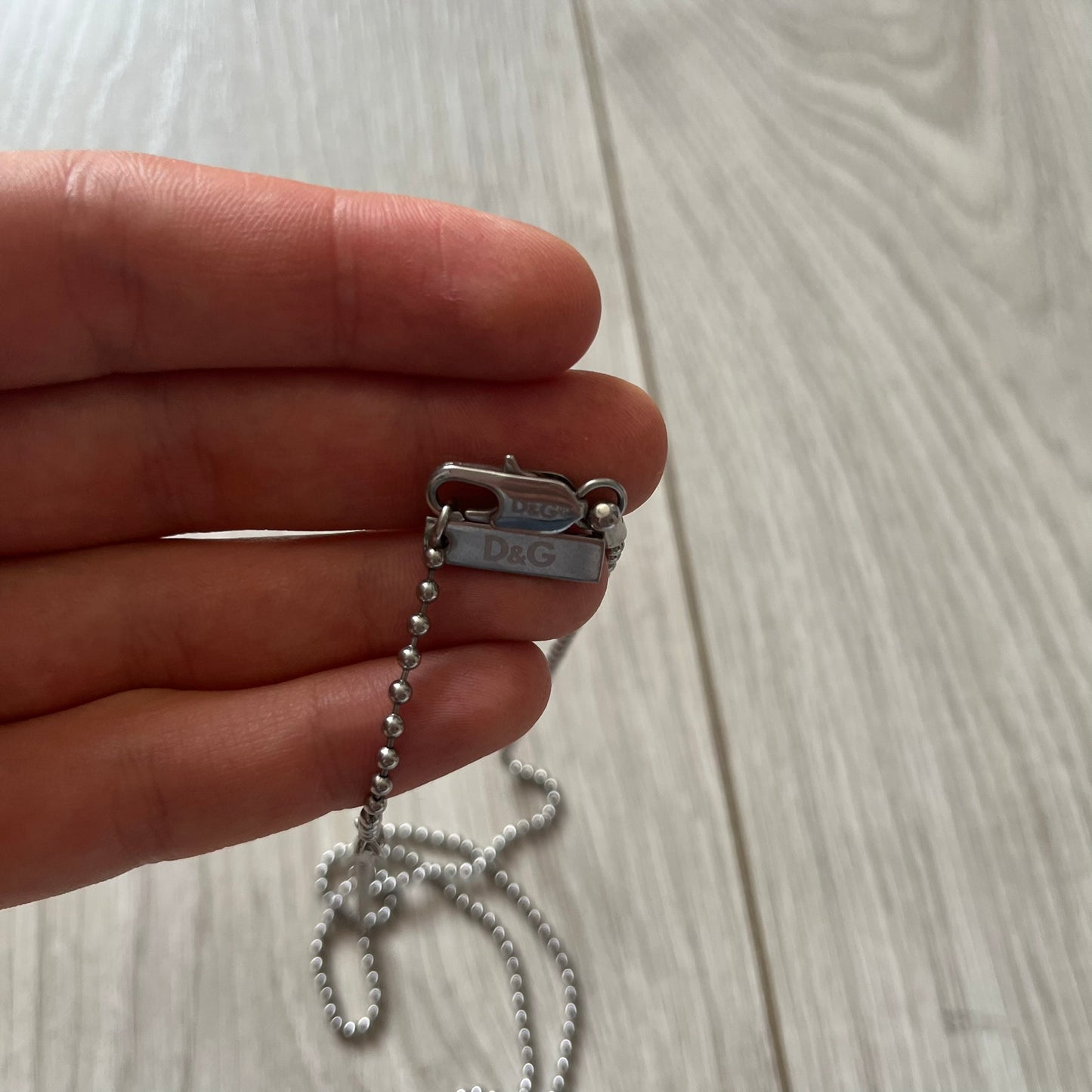 Hand holding a silver chain with a D&G tag on a light wooden surface