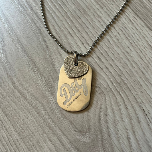 Dolce &amp; Gabbana pendant featuring a silver-tone dog tag engraved with the D&amp;G logo, topped with a separate rhinestone heart charm for a layered, eye-catching effect. Crafted from polished metal, it combines bold branding with a playful, sparkling detail.