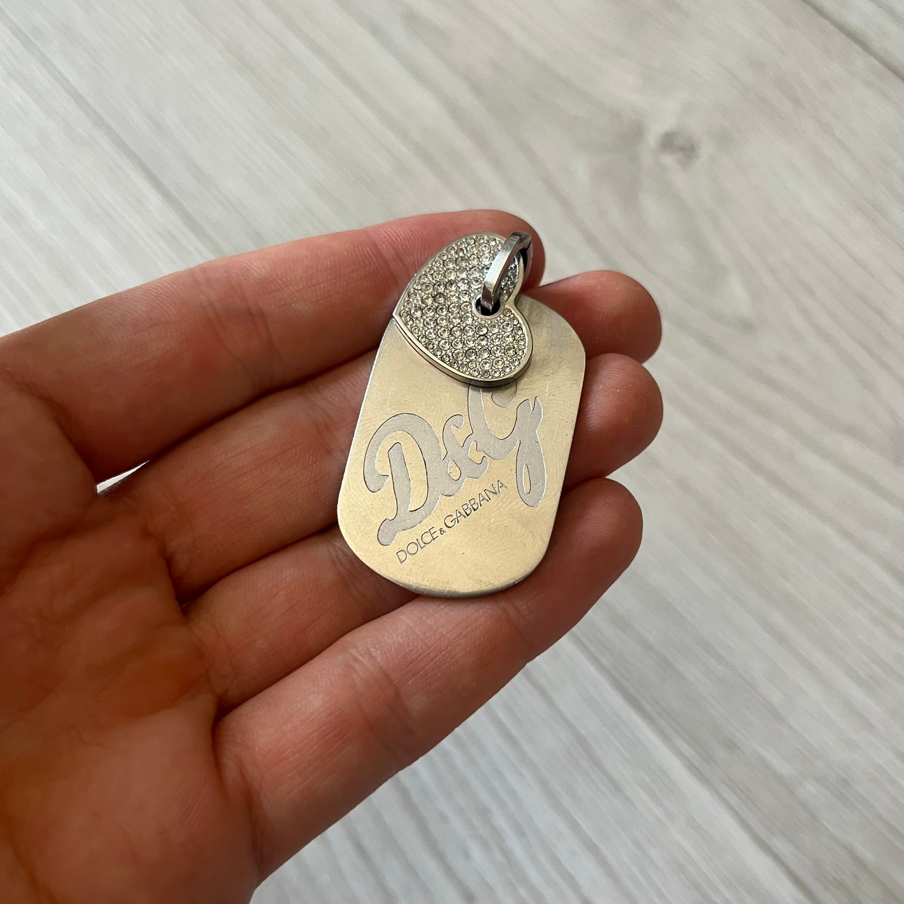 Dolce &amp; Gabbana pendant featuring a silver-tone dog tag engraved with the D&amp;G logo, topped with a separate rhinestone heart charm for a layered, eye-catching effect. Crafted from polished metal, it combines bold branding with a playful, sparkling detail.