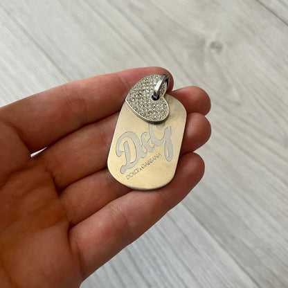 Dolce &amp; Gabbana pendant featuring a silver-tone dog tag engraved with the D&amp;G logo, topped with a separate rhinestone heart charm for a layered, eye-catching effect. Crafted from polished metal, it combines bold branding with a playful, sparkling detail.