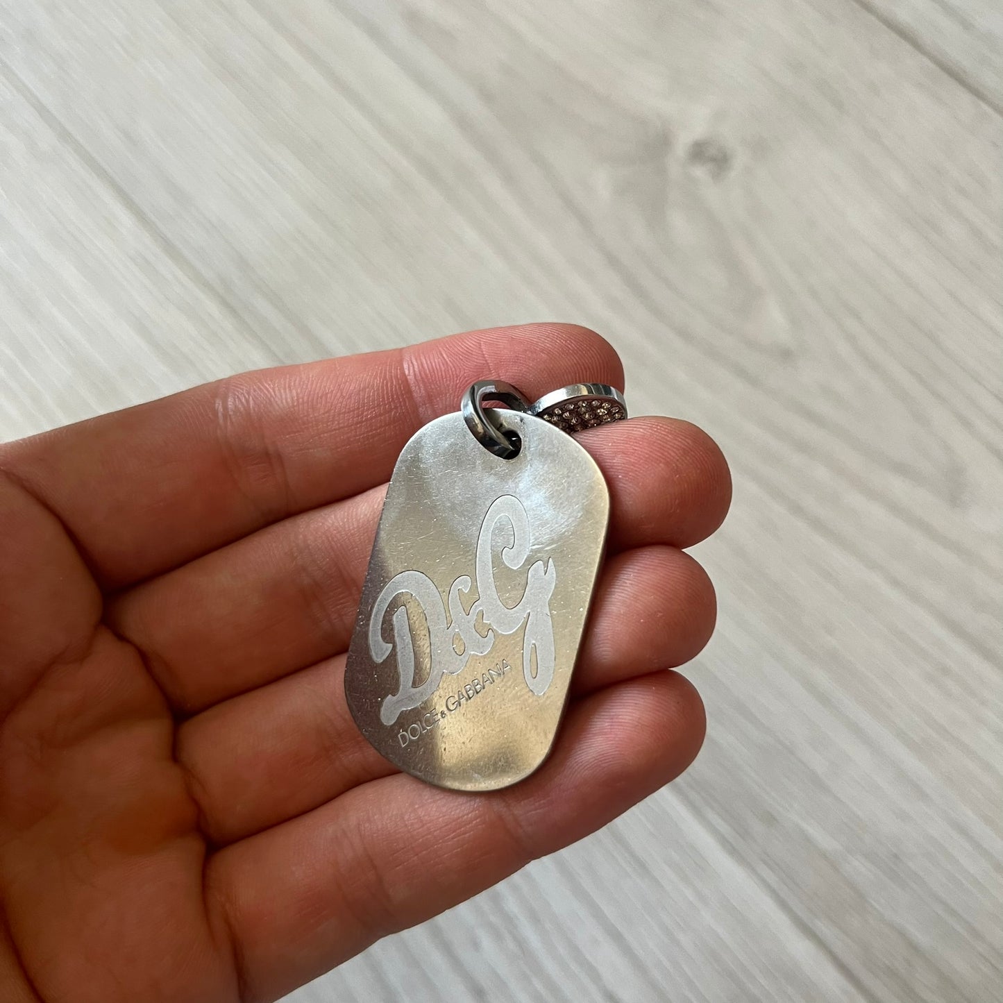Dolce &amp; Gabbana pendant featuring a silver-tone dog tag engraved with the D&amp;G logo, topped with a separate rhinestone heart charm for a layered, eye-catching effect. Crafted from polished metal, it combines bold branding with a playful, sparkling detail.