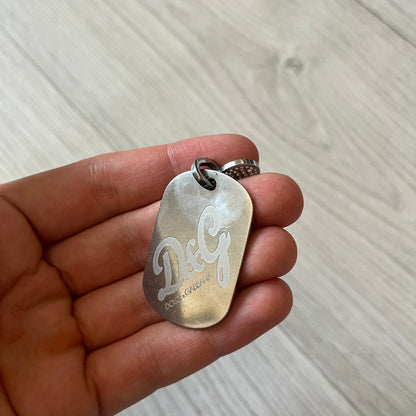 Dolce &amp; Gabbana pendant featuring a silver-tone dog tag engraved with the D&amp;G logo, topped with a separate rhinestone heart charm for a layered, eye-catching effect. Crafted from polished metal, it combines bold branding with a playful, sparkling detail.