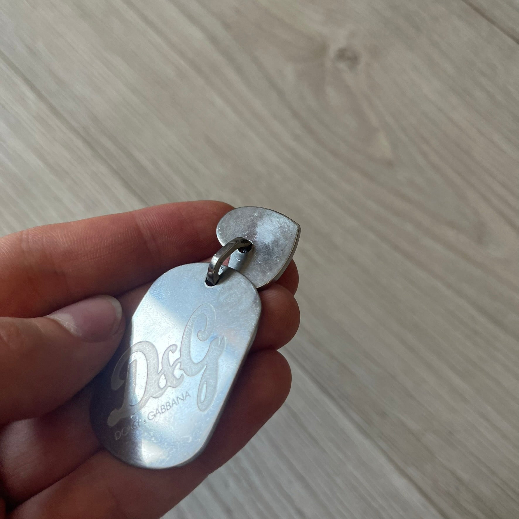 Dolce &amp; Gabbana pendant featuring a silver-tone dog tag engraved with the D&amp;G logo, topped with a separate rhinestone heart charm for a layered, eye-catching effect. Crafted from polished metal, it combines bold branding with a playful, sparkling detail.