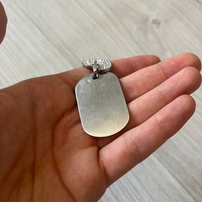 Hand holding a silver dog tag pendant on a light wooden surface
