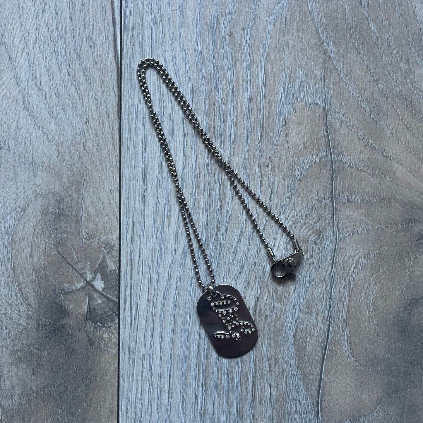 The Dolce & Gabbana Crystal Logo Dog Tag Necklace features a silver-tone ball chain with two pendants: a plain dog tag and a cutout D&G logo adorned with crystals. It has a large clasp engraved with D&G and model markings, from the 2000s.