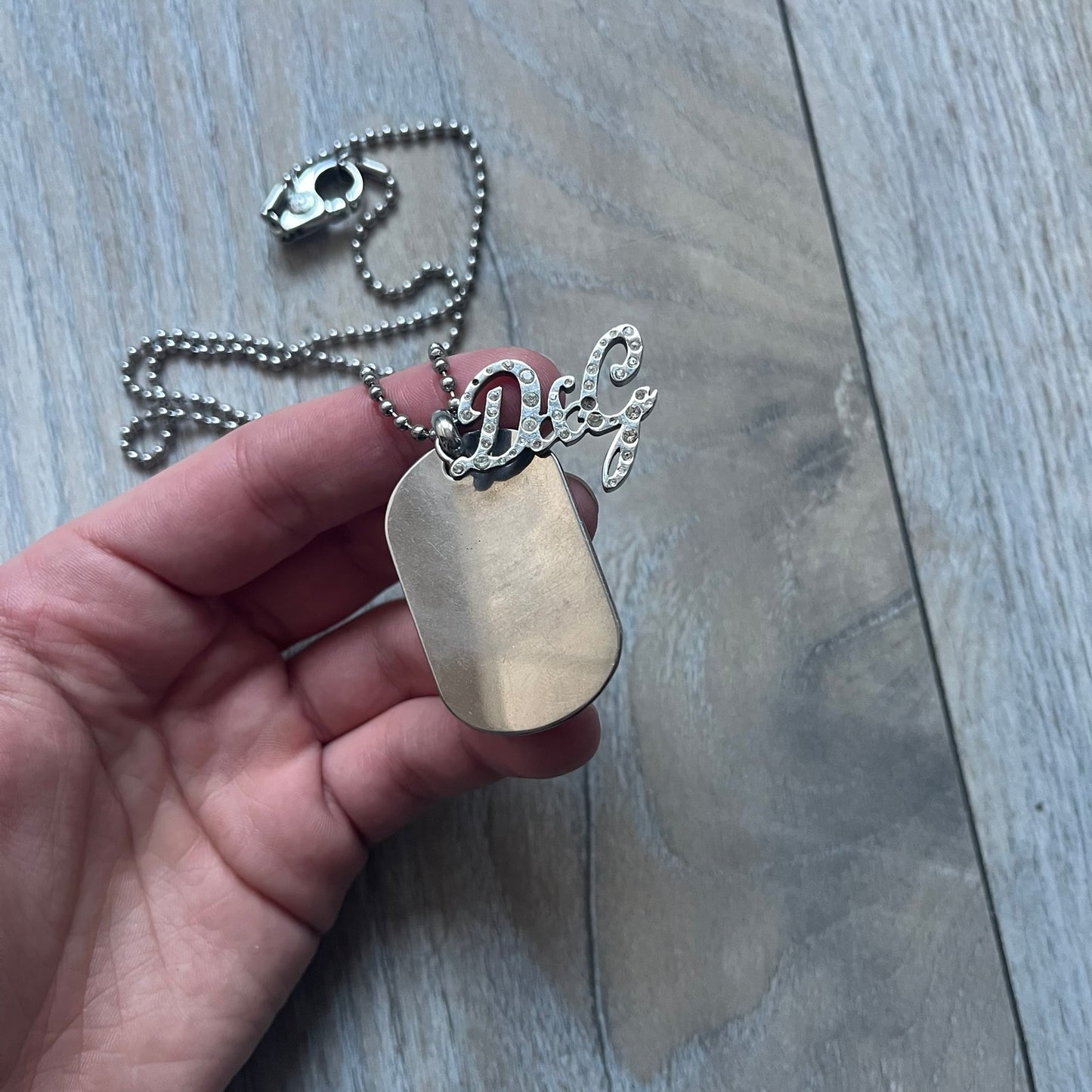 The Dolce & Gabbana Crystal Logo Dog Tag Necklace features a silver-tone ball chain with two pendants: a plain dog tag and a cutout D&G logo adorned with crystals. It has a large clasp engraved with D&G and model markings, from the 2000s.