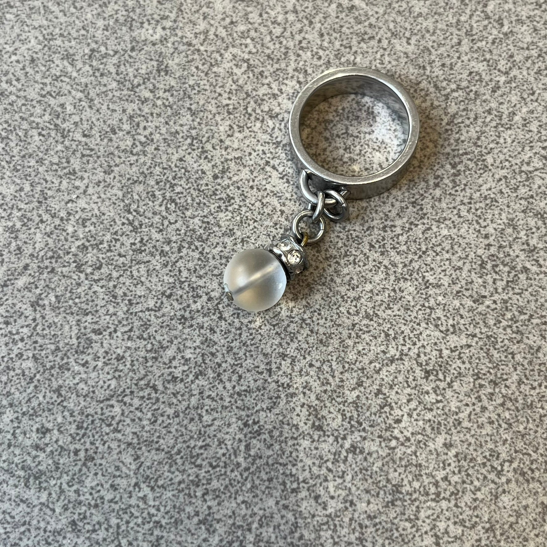 Dolce & Gabbana DJ0305 Pearl Charm Ring features a metal ring with a hanging charm consisting of a pearl with a crystal-like element above it. The charm is loose and moves freely from the ring. One additional charm is missing. The inside of the ring is engraved with a D&G logo.
