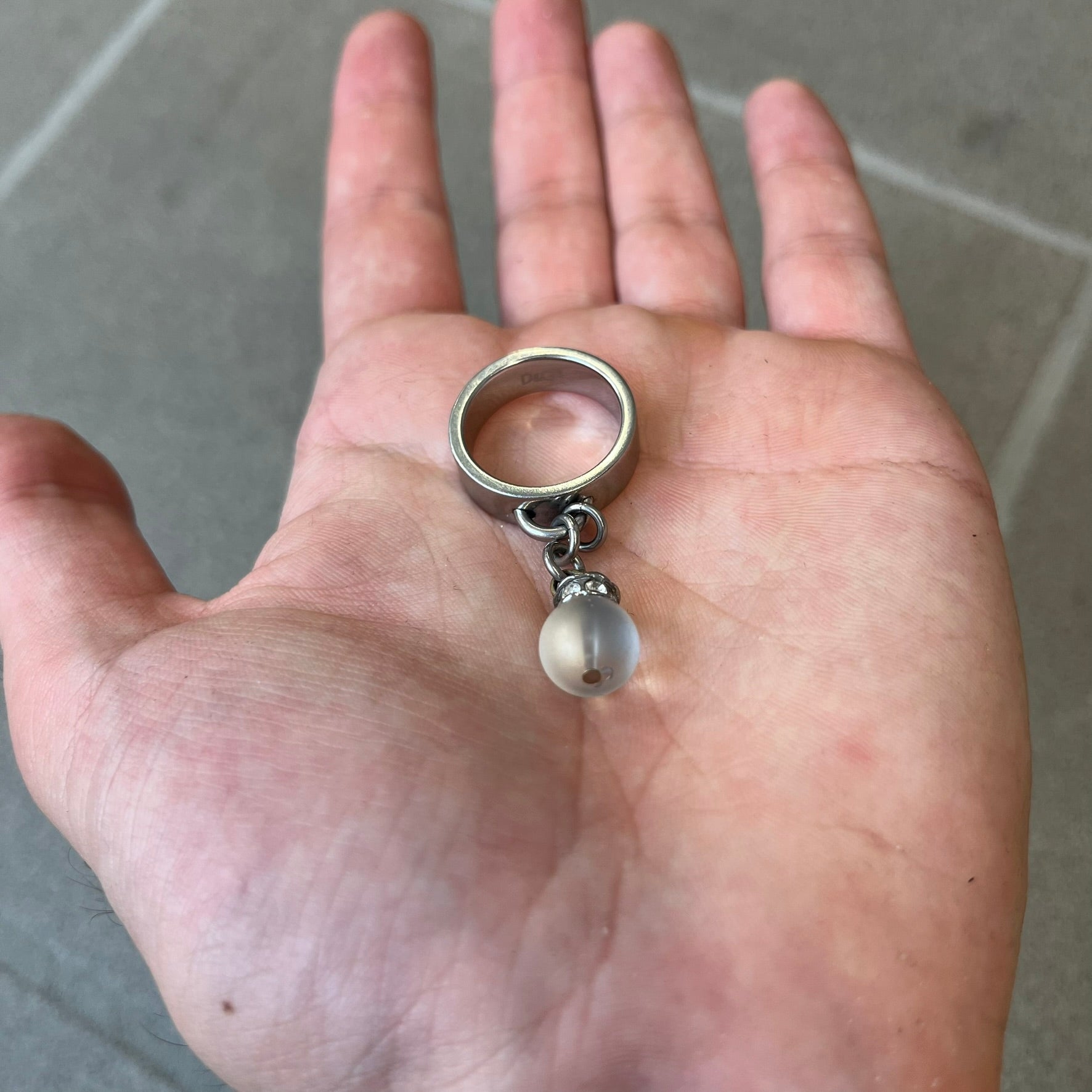 Dolce & Gabbana DJ0305 Pearl Charm Ring features a metal ring with a hanging charm consisting of a pearl with a crystal-like element above it. The charm is loose and moves freely from the ring. One additional charm is missing. The inside of the ring is engraved with a D&G logo.
