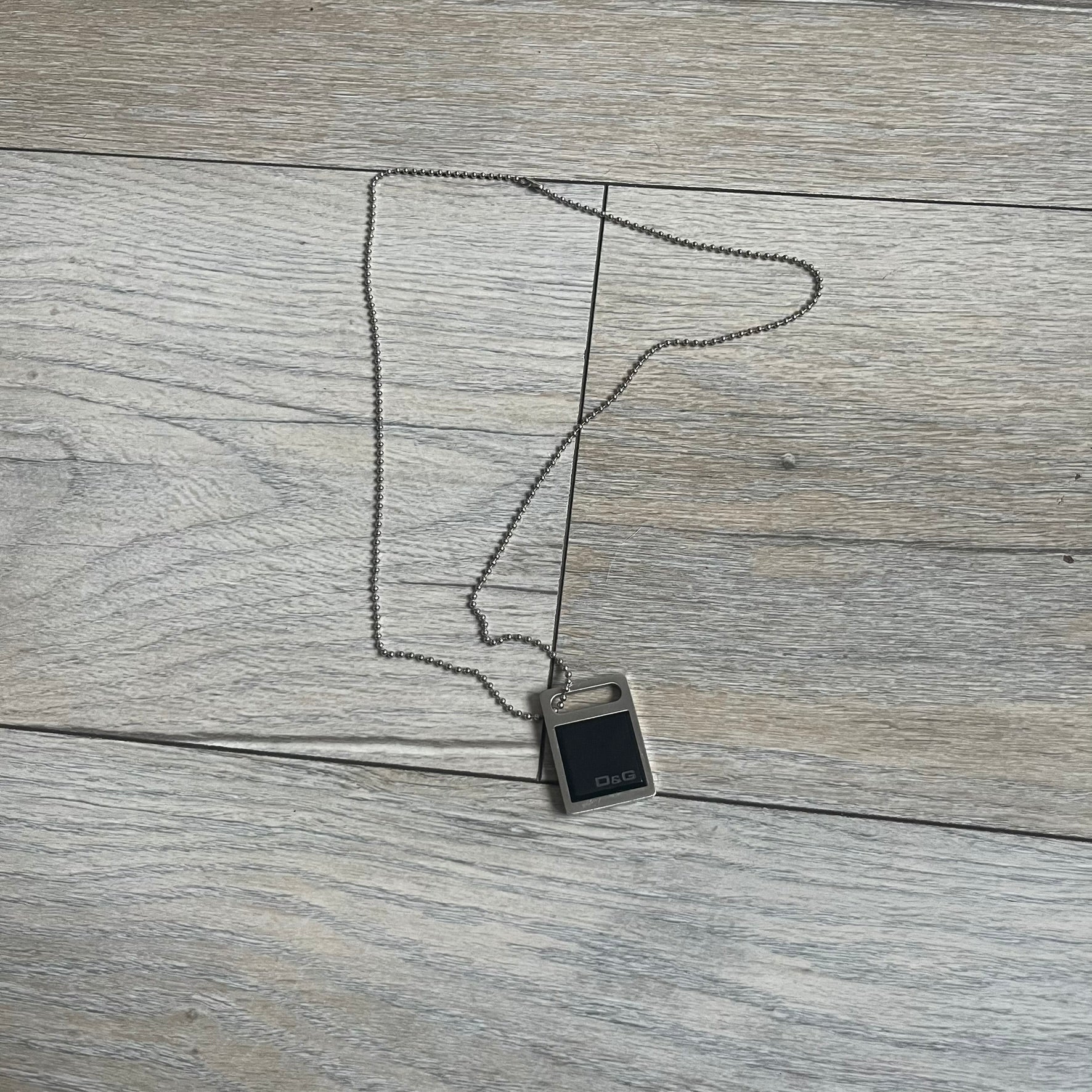 Dolce & Gabbana DJ0430 Black Stone Necklace is a 2000s necklace featuring a stainless steel pendant with a square black stone insert on the front. The lower right corner of the stone is engraved with a D&G logo. It comes with a steel ball chain.