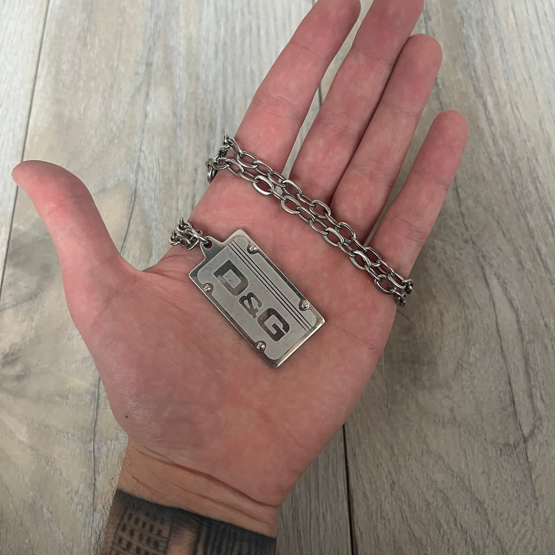 Dolce &amp; Gabbana Propaganda Dog Tag Necklace, featuring a silver stainless steel square tag with four visible screws and the D&amp;G logo engraved on the front. The tag hangs from a durable cable chain, combining sturdy construction with clean, industrial-inspired detailing.