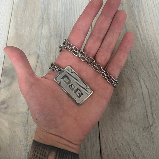 Dolce &amp; Gabbana Propaganda Dog Tag Necklace, featuring a silver stainless steel square tag with four visible screws and the D&amp;G logo engraved on the front. The tag hangs from a durable cable chain, combining sturdy construction with clean, industrial-inspired detailing.