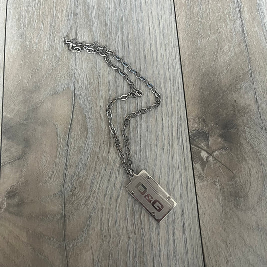 Dolce &amp; Gabbana Propaganda Dog Tag Necklace, featuring a silver stainless steel square tag with four visible screws and the D&amp;G logo engraved on the front. The tag hangs from a durable cable chain, combining sturdy construction with clean, industrial-inspired detailing.