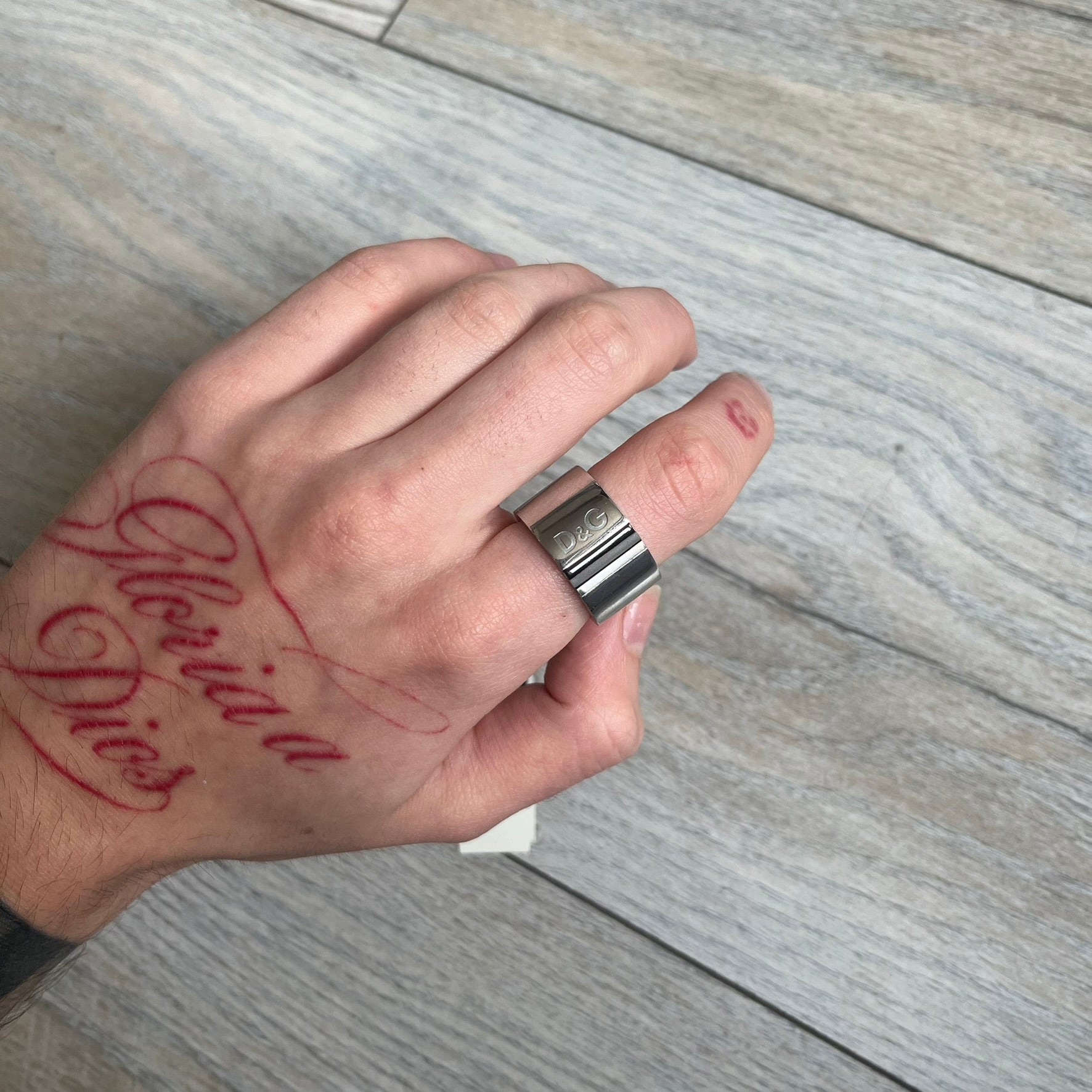 Dolce & Gabbana DJ0551 3 Lines Ring is a stainless steel ring featuring an engraved D&G logo placed vertically along the band. Next to the logo are three parallel lines in black, grey, and white. The design is minimal with a smooth metal finish.