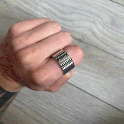 Dolce & Gabbana DJ0551 3 Lines Ring is a stainless steel ring featuring an engraved D&G logo placed vertically along the band. Next to the logo are three parallel lines in black, grey, and white. The design is minimal with a smooth metal finish.