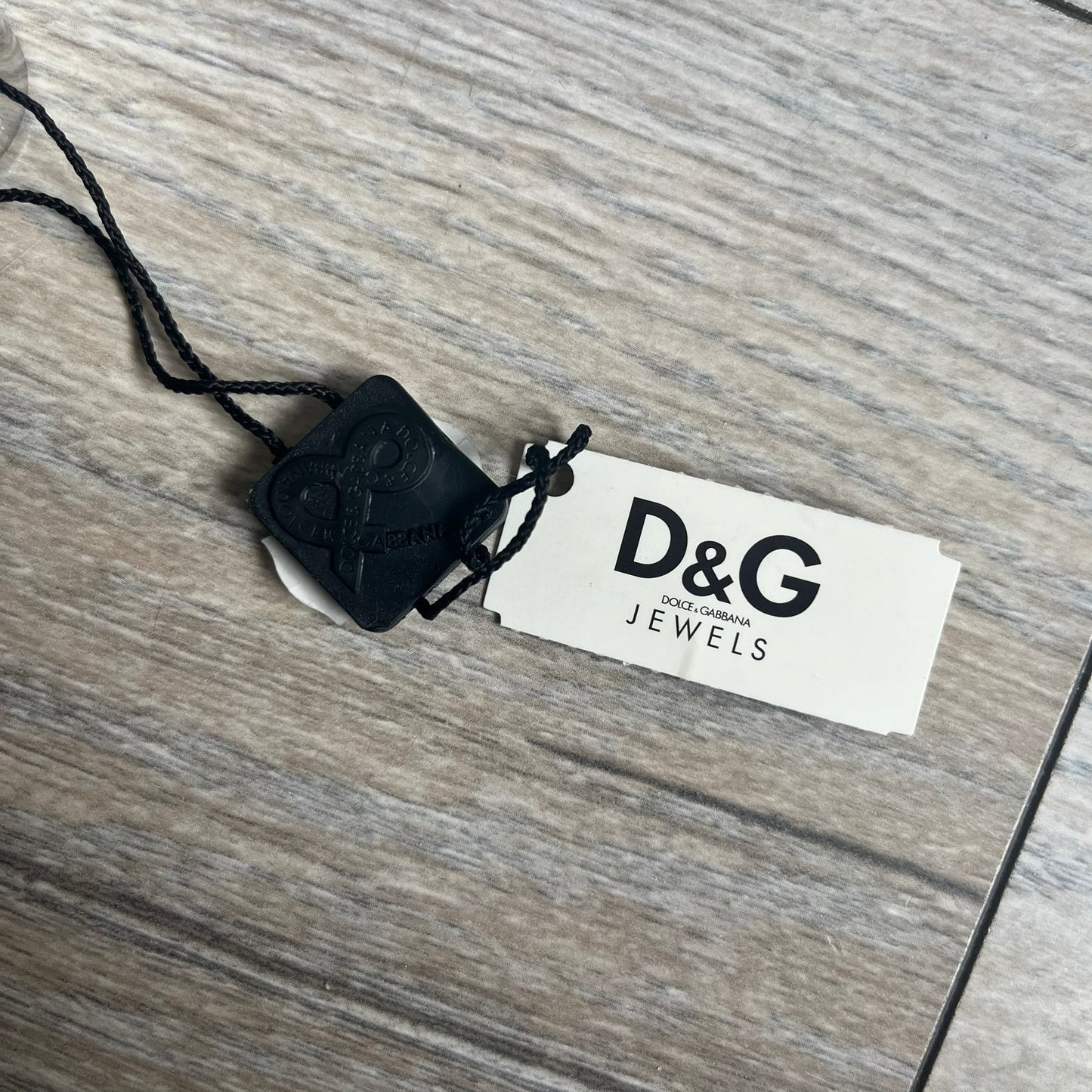 Dolce & Gabbana DJ0551 3 Lines Ring is a stainless steel ring featuring an engraved D&G logo placed vertically along the band. Next to the logo are three parallel lines in black, grey, and white. The design is minimal with a smooth metal finish.