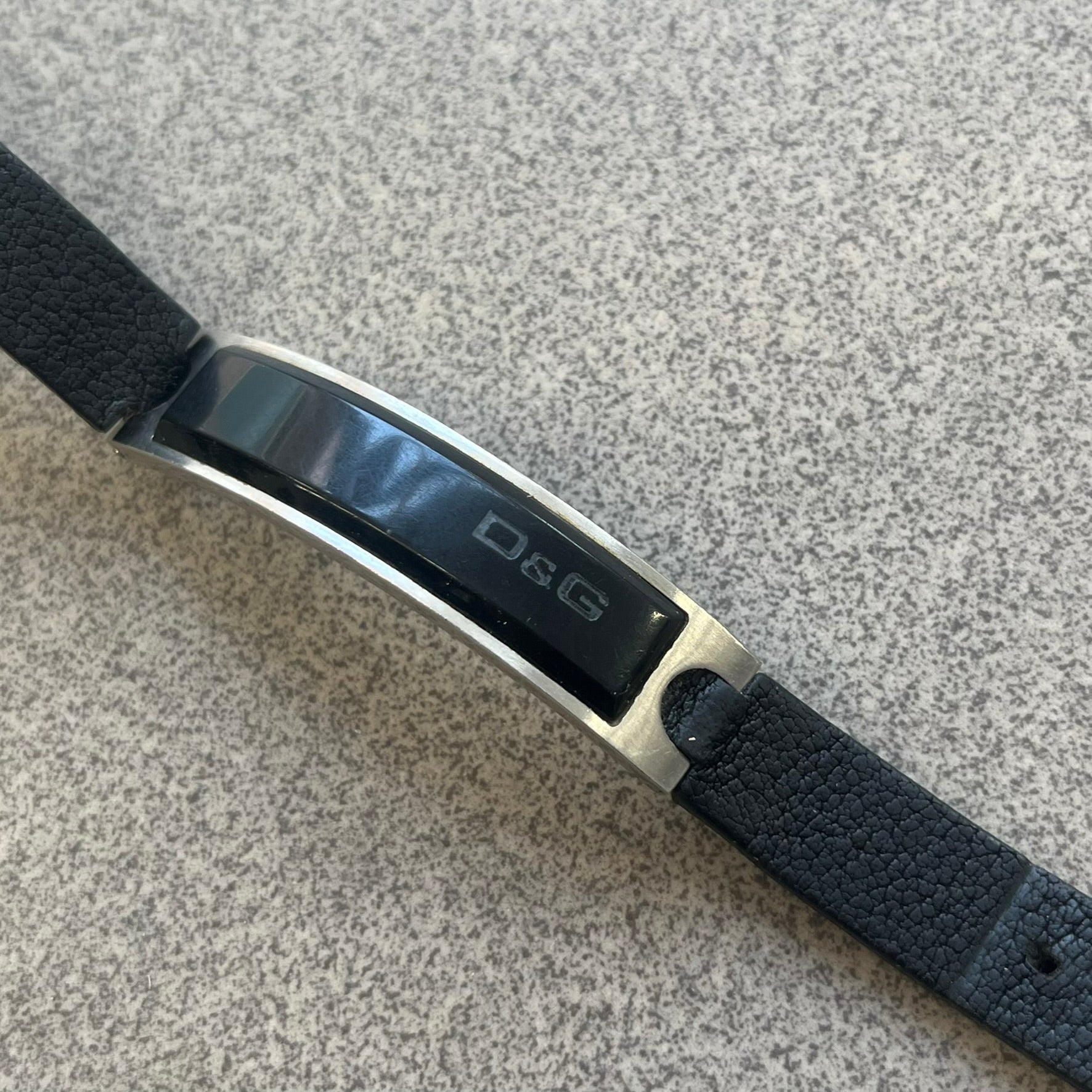 Dolce & Gabbana DJ0574 Black Stone Bracelet is a black leather bracelet featuring a stainless steel central plaque. The plaque includes a rectangular black stone detail with an engraved D&G logo on the right side. The clasp is also engraved with the D&G logo, completing the minimal design.
