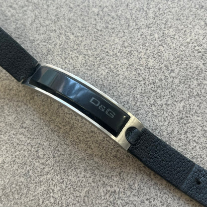 Dolce & Gabbana DJ0574 Black Stone Bracelet is a black leather bracelet featuring a stainless steel central plaque. The plaque includes a rectangular black stone detail with an engraved D&G logo on the right side. The clasp is also engraved with the D&G logo, completing the minimal design.
