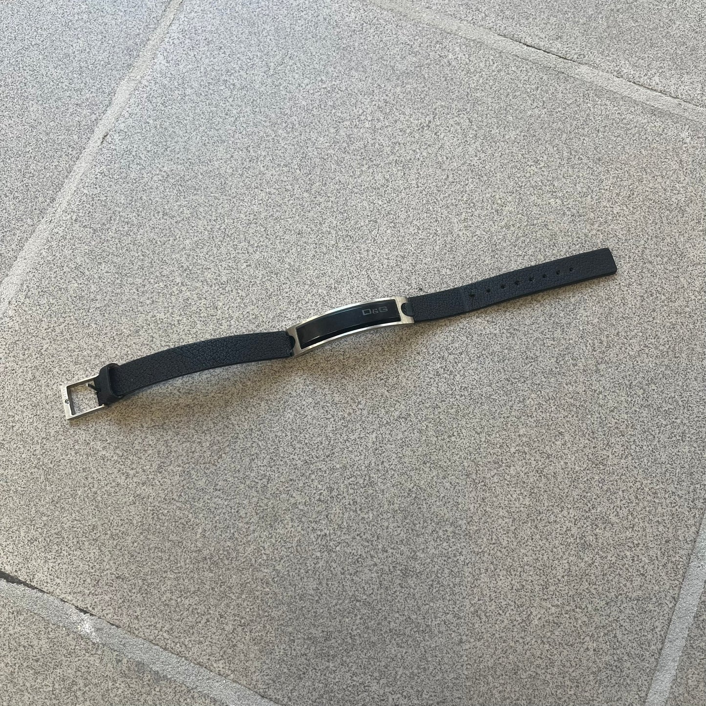 Dolce & Gabbana DJ0574 Black Stone Bracelet is a black leather bracelet featuring a stainless steel central plaque. The plaque includes a rectangular black stone detail with an engraved D&G logo on the right side. The clasp is also engraved with the D&G logo, completing the minimal design.
