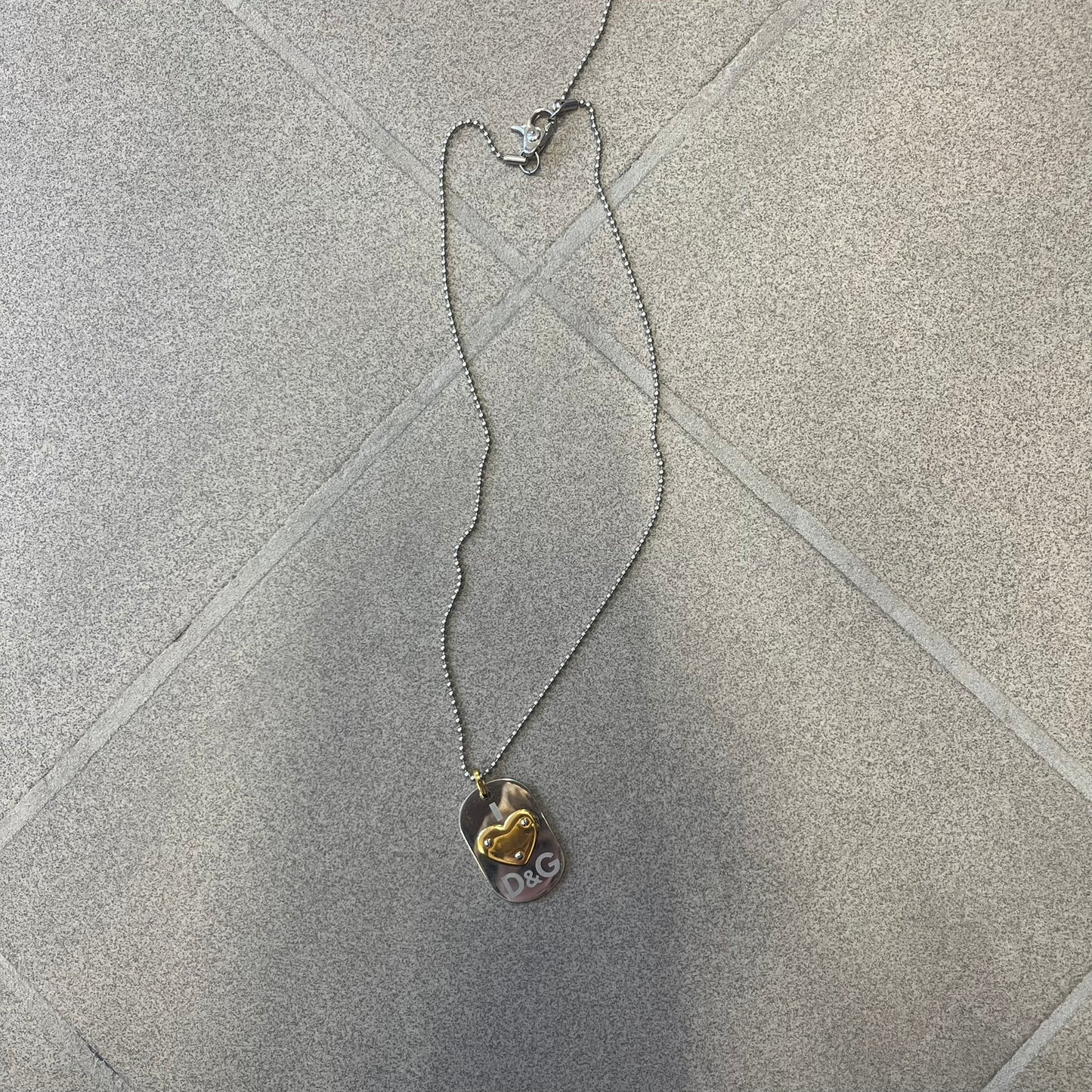 Dolce & Gabbana DJ0637 Gold Heart Necklace features a silver-tone stainless steel ball chain with a dog tag-style pendant. The pendant is engraved with a subtle "I" above a gold-tone heart secured by three visible screws, with "D&G" engraved below to form the "I ❤️ D&G" design. The back of the pendant has a "K.O" engraving from a previous owner. Comes with the original dust bag.
