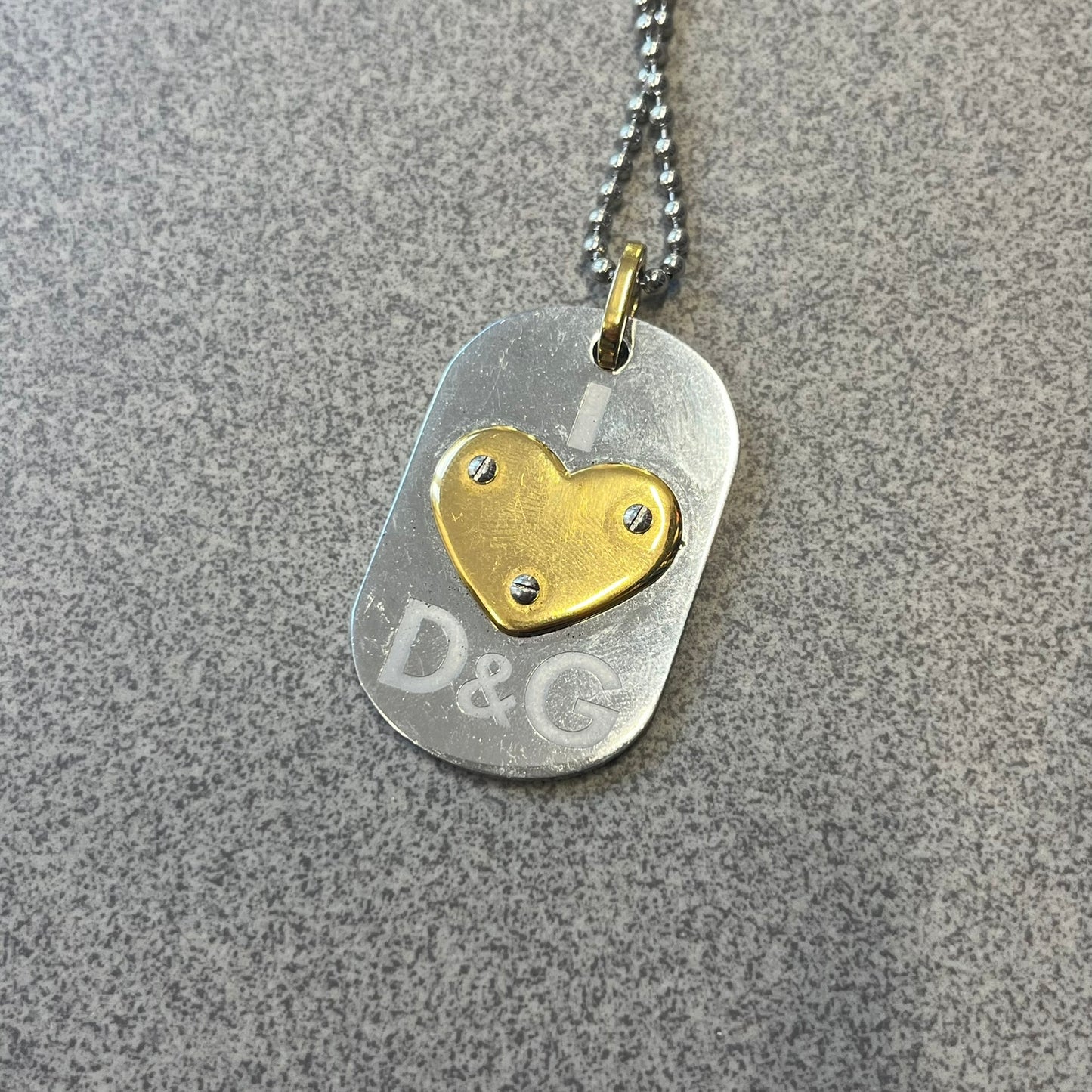 Dolce & Gabbana DJ0637 Gold Heart Necklace features a silver-tone stainless steel ball chain with a dog tag-style pendant. The pendant is engraved with a subtle "I" above a gold-tone heart secured by three visible screws, with "D&G" engraved below to form the "I ❤️ D&G" design. The back of the pendant has a "K.O" engraving from a previous owner. Comes with the original dust bag.
