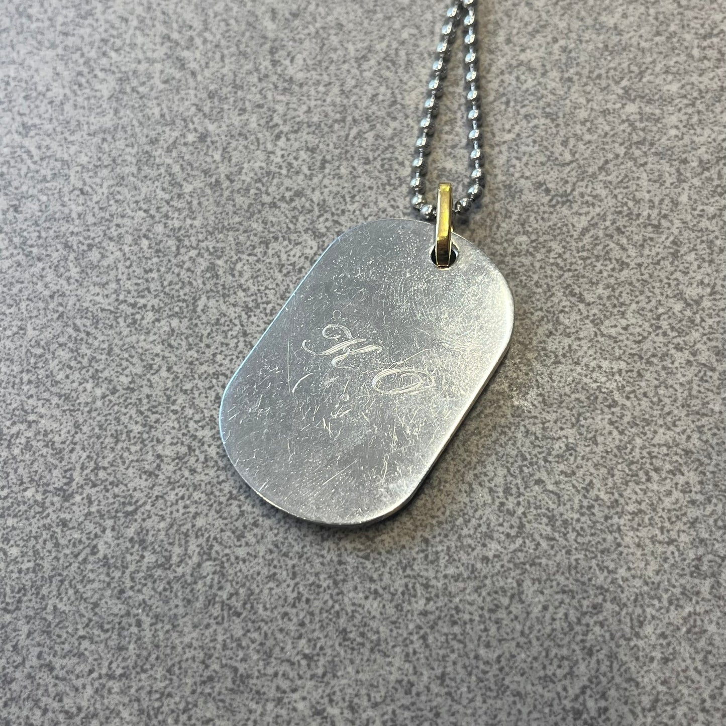 Dolce & Gabbana DJ0637 Gold Heart Necklace features a silver-tone stainless steel ball chain with a dog tag-style pendant. The pendant is engraved with a subtle "I" above a gold-tone heart secured by three visible screws, with "D&G" engraved below to form the "I ❤️ D&G" design. The back of the pendant has a "K.O" engraving from a previous owner. Comes with the original dust bag.
